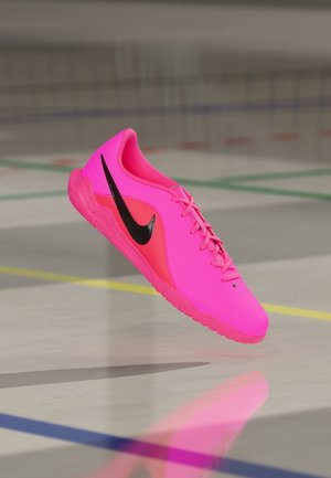 Chaussure de football d'intérieur Nike rose vif avec logo swoosh noir, flottant au-dessus d'un sol de gymnase marqué de lignes colorées.