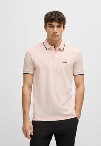 Polo shirt rosa chiaro realizzato in tessuto liscio, con una rifinitura nera sul colletto e sulle maniche, e un piccolo logo nero sul petto.
