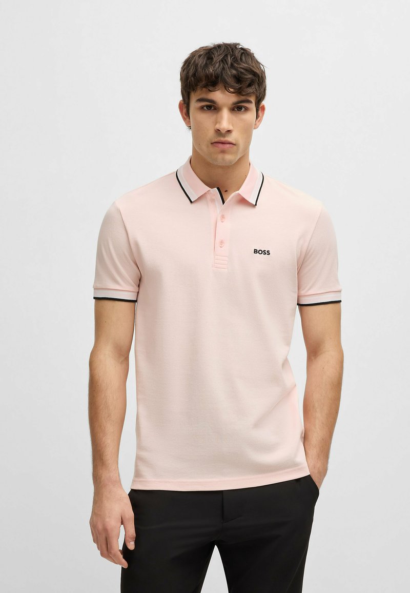 Polo shirt rosa chiaro realizzato in tessuto liscio, con una rifinitura nera sul colletto e sulle maniche, e un piccolo logo nero sul petto.