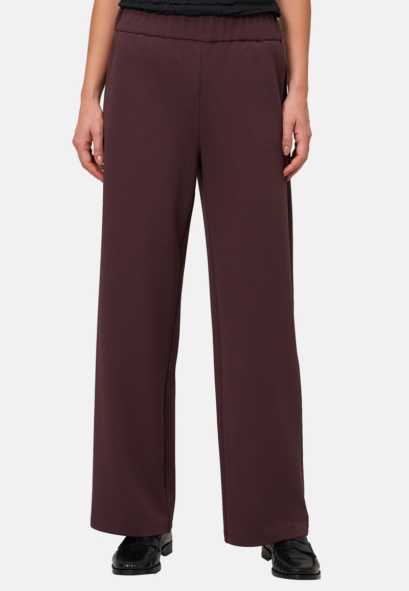 Vrouw in donk paarse wijde broek en zwarte loafers, staand tegen een eenvoudige lichte achtergrond.