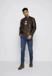 Veste en cuir marron de style aviateur sur un pull en maille à motifs, associée à un jean bleu et des bottes à lacets marron, se tenant devant un fond neutre.