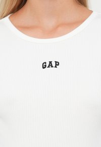 Witte geribbelde T-shirt met een ronde halslijn, voorzien van zwarte "GAP" borduring op de borst. De stof is zacht en iets rekbaar.