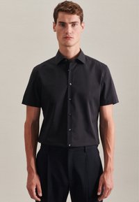 Maglietta nera a maniche corte in tessuto liscio, con colletto e placket con bottoni scuri. Il modello indossa pantaloni scuri formali.