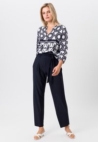 Blouse à motifs noir et blanc avec un col en V et des manches 3/4, associée à un pantalon taille haute en tissu navy et des sandales à talons blancs.