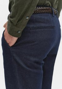 Ciemne dżinsowe spodnie o gładkiej fakturze, z tylną kieszenią i plecionym brązowym paskiem. Koszula w kolorze oliwkowym z zapinanymi mankietami.