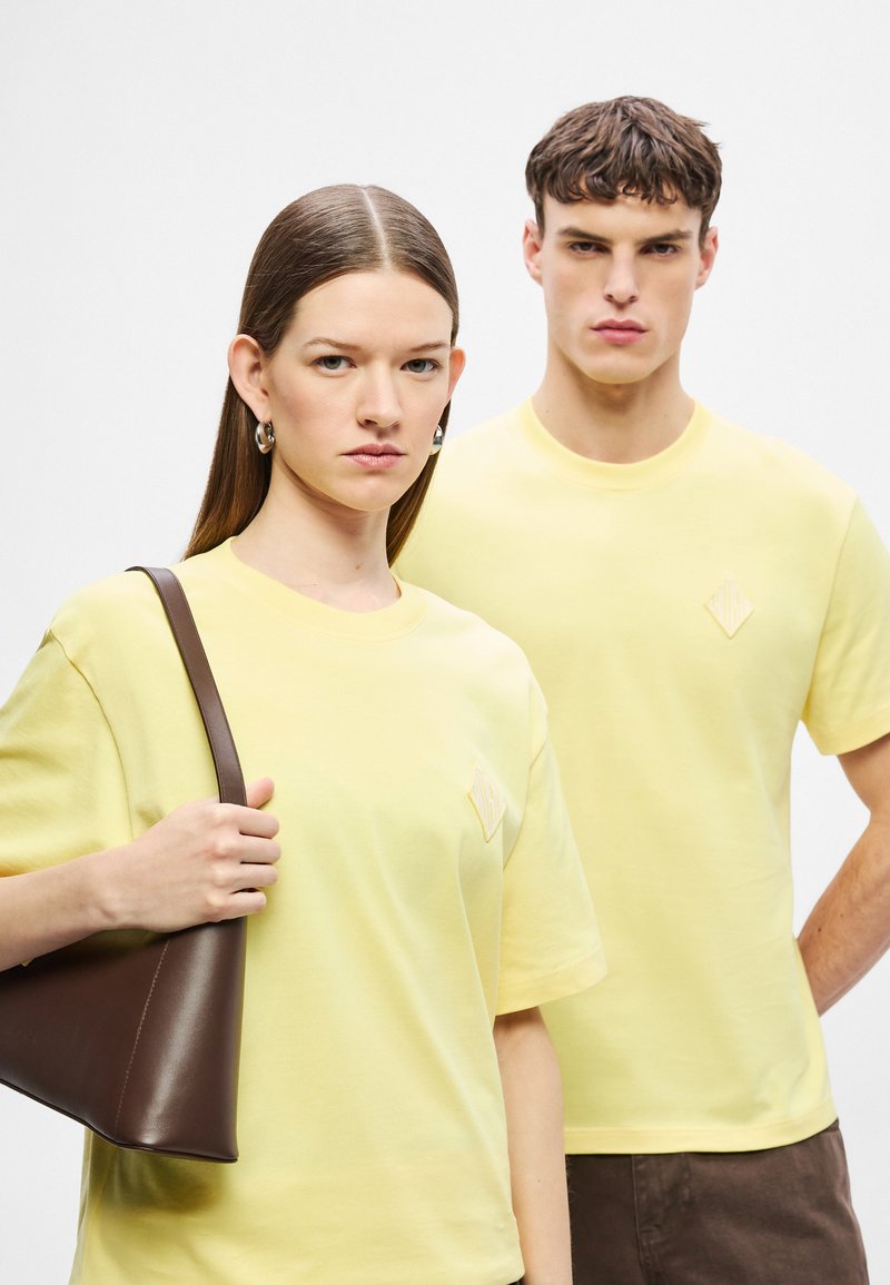 Le T-shirt gialle in cotone presentano una toppa con logo a forma di diamante, abbinati a una borsa in pelle marrone. I capelli della modella sono lisci e diritti.