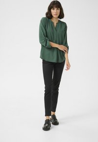 Blusa verde bosco con maniche a tre quarti a doppio risvolto, scollatura a V e una vestibilità rilassata, abbinata a pantaloni neri attillati e scarpe nere.