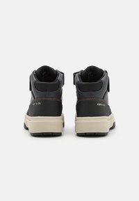 Chaussures noires et grises montantes en matériau synthétique avec une texture lisse, dotées d'une semelle en caoutchouc beige et d'une bride ajustable.