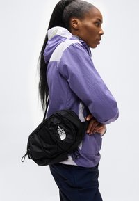 Άτομο με μωβ και λευκό μπουφάν που φοράει μαύρη τσάντα ώμου The North Face, στέκεται πλάγια με τα χέρια σταυρωμένα, μπροστά σε απλό φόντο.