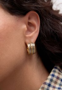 Gold-tone hoop earrings featuring multiple stacked, rounded bands with a polished finish, worn close to the ear against short hair.