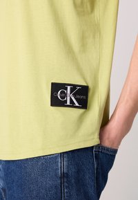 Világoszöld pamut póló, fekete téglalap alakú logóval, amelyen fehér szöveg olvasható: "Calvin Klein Jeans" és egy 'CK' embléma.
