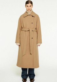 Lang, beige uldblandingsfrakke med en bred krave, bælte og knaplukning. Stylet over en beige rullekravetrøje og mørkeblå brede jeans.