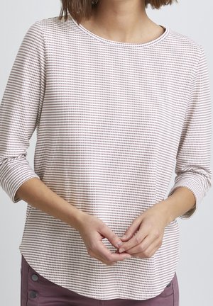 Kobieta w beżowo-białej koszuli w paski z długim rękawem, w mauve spodniach, delikatnie splecione dłonie na wysokości talii na neutralnym tle.