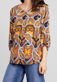 Kvinde iført bluse med paisley-mønster i gul, orange, marineblå og hvid med trekvart ærmer sammen med blå jeans.