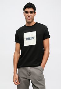 Fekete, kerek nyakú póló felhajtott ujjakkal. A mellkason bézs és kék négyzet alakú grafikával díszített logó található, pamut anyagból készült.