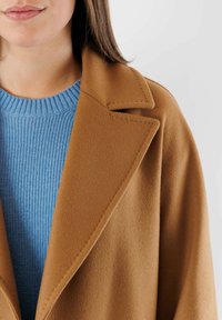 Cappotto di lana color cammello con rever a punta, abbinato a un maglione di maglia blu testurizzato. Primo piano che mette in evidenza i dettagli del tessuto e delle cuciture.