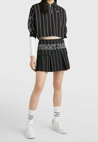 Zwart-wit gestreepte cropped polo met rits, gecombineerd met een bijpassende geplooide rok met de tekst "TOMMY JEANS". Witte sokken en sneakers maken de look af.