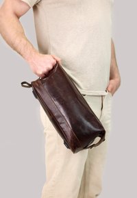 Brown leather bag with a textured finish, featuring a side handle and a woven accent, held by a person in light-colored clothing.