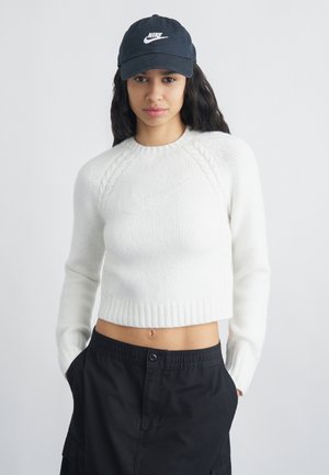 Joven con el cabello largo y oscuro, lleva una gorra negra de Nike, un suéter corto blanco y pantalones negros, de pie con las manos en los bolsillos.