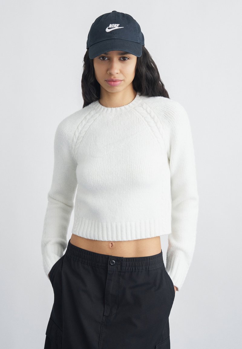 Νεαρή γυναίκα με μακριά σκούρα μαλλιά, φορώντας μαύρο καπέλο Nike, λευκό κοντό πουλόβερ και μαύρο παντελόνι, στέκεται με τα χέρια στις τσέπες.