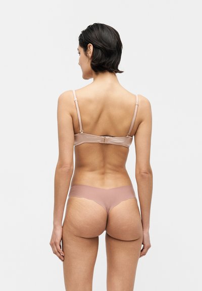 Frau mit kurzen dunklen Haaren, die einen beigen BH und einen passenden Tanga trägt, steht mit dem Rücken zu einem einfarbigen hellen Hintergrund.