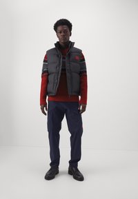 Zwarte puffervest met hoge kraag over een rode trui met een geometrisch patroon. Marineblauwe cargobroek; zwarte schoenen. Simpel, casual outfit.