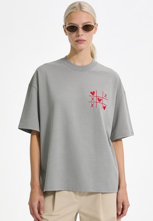 Vrouw met blond haar die een zonnebril draagt en een los grijs T-shirt met een rood boter-kaas-en-eieren hart en X-ontwerp op de borst.