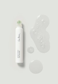 Un tube blanc de sérum pour les yeux à l'eau de raisin avec un roll-on vert, accompagné de gouttes de gel transparent sur un fond gris clair.