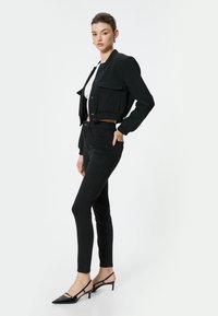 Chaqueta negra corta con dos bolsillos frontales, combinada con jeans ajustados negros y tacones de punta. Tejido suave con un diseño ajustado.