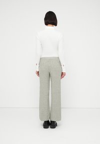 Personne aux cheveux courts et foncés, portant un haut blanc à manches longues et un pantalon gris large en maille torsadée, debout face à un fond blanc uni.