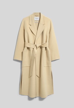 Kvinde med langt rødt hår iført en beige lang frakke over en beige top og vide blå jeans, stående mod en ensfarvet baggrund.