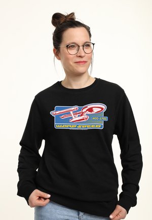 Kvinde med briller og hår i en knold iført en sort sweatshirt med et rødt Starship Enterprise-tryk og teksten "Warp Speed NCC-1701".