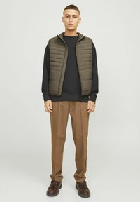 Ärmelleste olivfarbene Steppweste über einem schwarzen Sweatshirt, kombiniert mit braunen, gefalteten Hosen und dunkelbraunen Schuhen. Schlichtes Design und strukturierte Passform.