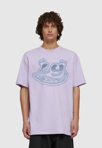 Svetle fialové bavlněné tričko s krátkým rukávem, s velkým modrým grafickým designem rakety a hvězd na přední straně.