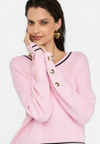 Helles rosa Sweatshirt mit schwarzen Streifenakzenten, versehen mit goldenen Knöpfen an den Bündchen. Das Material wirkt weich und hat eine glatte Textur.