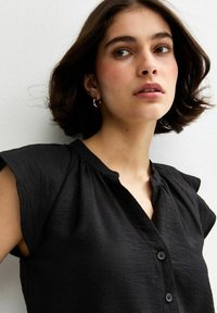 Camisa negra de cuello con mangas cortas tipo cap sleeve, con un tejido texturizado y una botonera en la parte delantera. Diseño minimalista.
