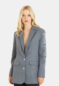 Blazer grigio su misura con scollo a V profondo, due bottoni e ricami dettagliati sulle maniche. Realizzato in un tessuto testurizzato con alette per le tasche.
