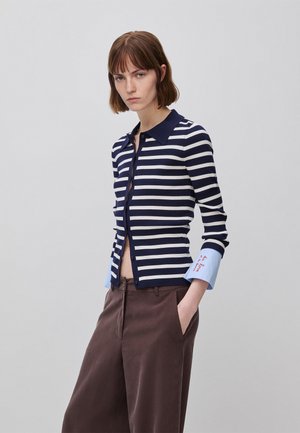 Frau mit kurzen braunen Haaren, die ein marineblau-weiß gestreiftes Langarmshirt mit blauen Bündchen trägt, auf denen roter Text gestickt ist, und braune Hosen mit weitem Bein.