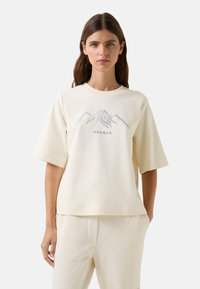 Lys cremefarvet t-shirt med korte ærmer lavet af blødt stof, med et bjergmotiv og mærkenavnet "BOGNER" broderet i grå.