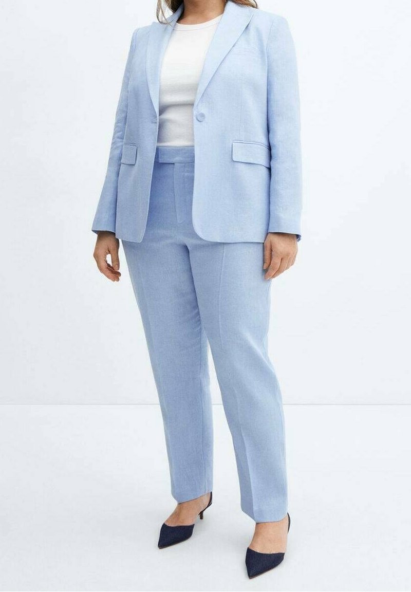 Personne portant un costume bleu clair avec un blazer et un pantalon, une chemise blanche et des talons noirs pointus, debout sur un fond blanc.