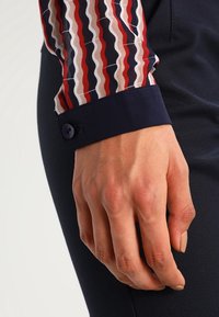 Camicia a maniche lunghe colorata con motivo ondulato rosso, bianco e blu navy, con un polsino blu navy scuro e dettaglio a bottone. Mano appoggiata su una superficie scura.