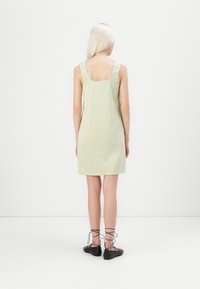 Vestido em gingham verde claro com costas quadradas e alças largas, até aos joelhos. Usado com sapatos pretos de amarrar nas pernas à mostra.