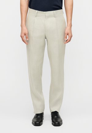 Prírodné béžové nohavice zo svetlého ľanu s rovnakým strihom, nosené s čiernymi formálnymi šnúrkami a čiernym zastrčeným topom, zobrazené od pása po nohy.