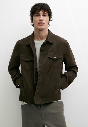 Jeune homme aux cheveux foncés portant une veste marron boutonnée sur une chemise gris clair et un pantalon gris, debout les mains dans les poches.