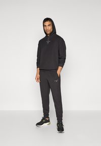 Schwarzer Hoodie mit Kordelzug und Logo, kombiniert mit passenden Jogginghosen. Getragen mit schwarzen Sportschuhen mit grünen Akzenten.