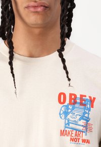 Beige cotton t-shirt featuring a graphic in red and blue with text "OBEY MAKE ART NOT WAR" and a design of a printing press.