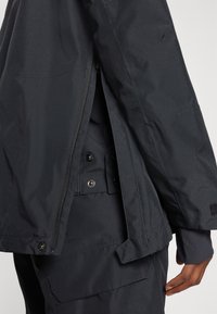 Černá nepromokavá bunda s zapínáním na zip, nastavitelnými manžetami a patentkami, hladkou matnou texturou a minimalistickým designem.