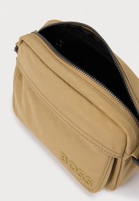 Μπεζ υφασμάτινη τσάντα ταχυδρόμου με μαύρο εσωτερικό, κλείσιμο με φερμουάρ, ανάγλυφο λογότυπο στο μπροστινό μέρος και λεία υφή.