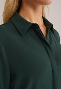 Detailní záběr osoby s tmavě zelenou košilí s límečkem, zobrazující výstřih, límeček a část oblasti ramen.