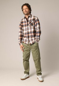 Flanellen shirt in bruin, wit en marineblauw ruitpatroon; heeft een enkele borstzak. Gepaard met olijfgroene broek en witte sneakers.